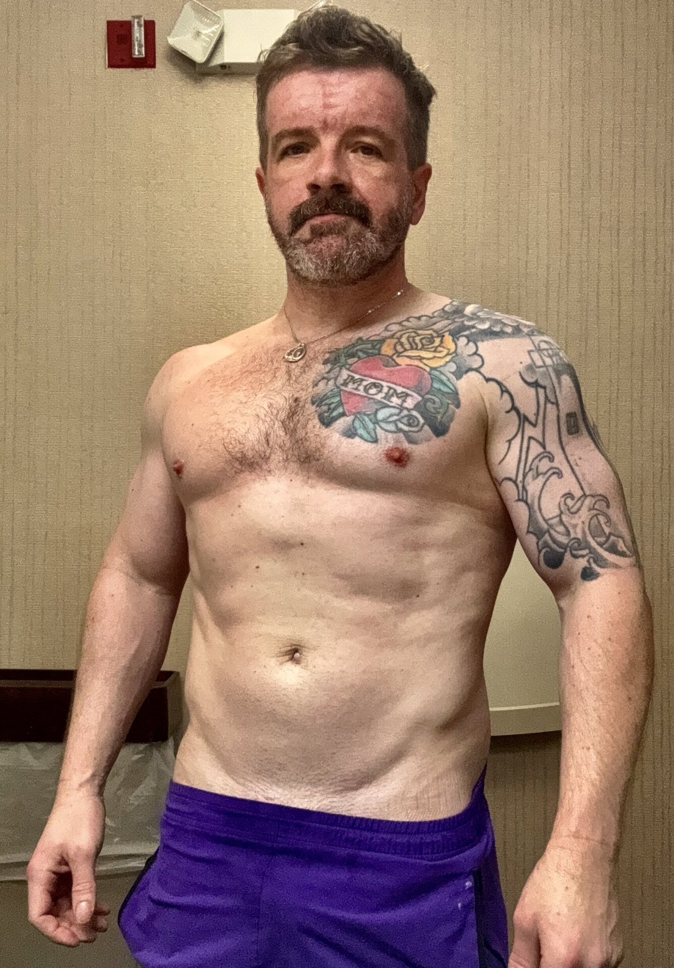Billy Pilgrim portrait — shirtless, salt-and-pepper beard, chest tattoo with 'MOM' heart banner and rose, full sleeve on left arm, gold pendant chain, purple shorts