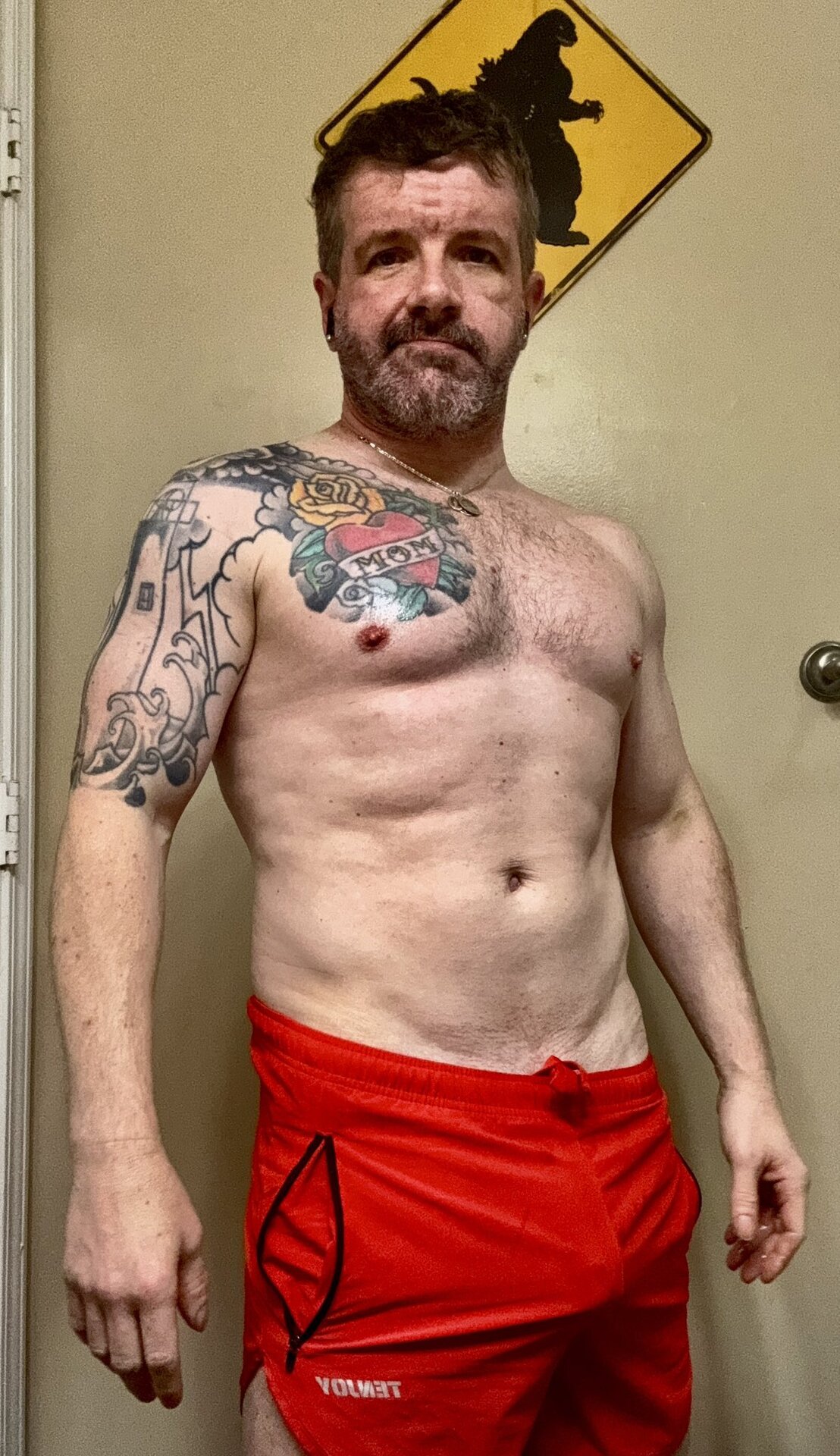 Billy Pilgrim in front of a Godzilla road-sign — shirtless, red FENDY shorts, tattoo sleeve visible, hotel-room setting