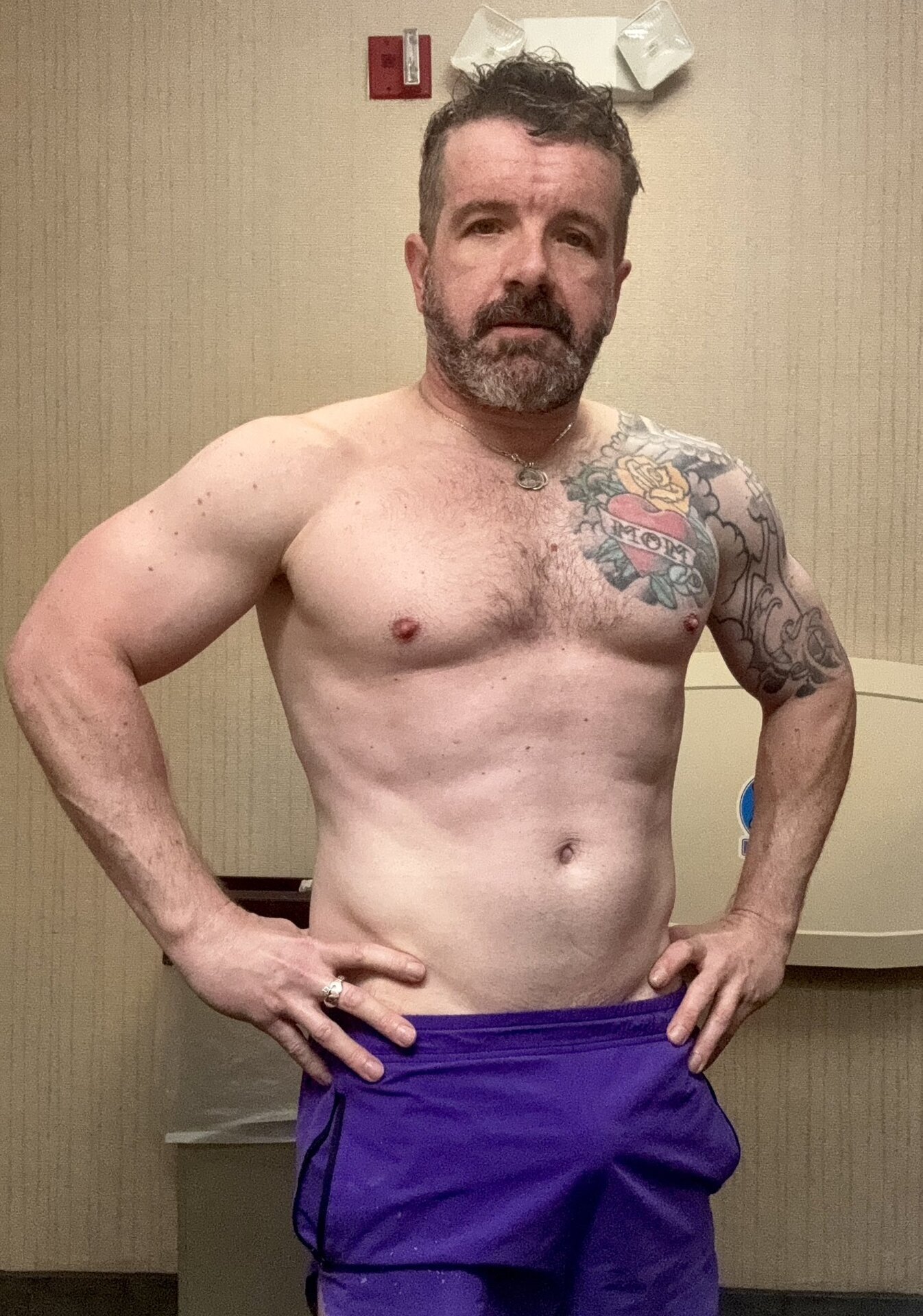 Billy Pilgrim hands-on-hips pose — shirtless, full salt-and-pepper beard, chest tattoos visible, confident stance, purple shorts