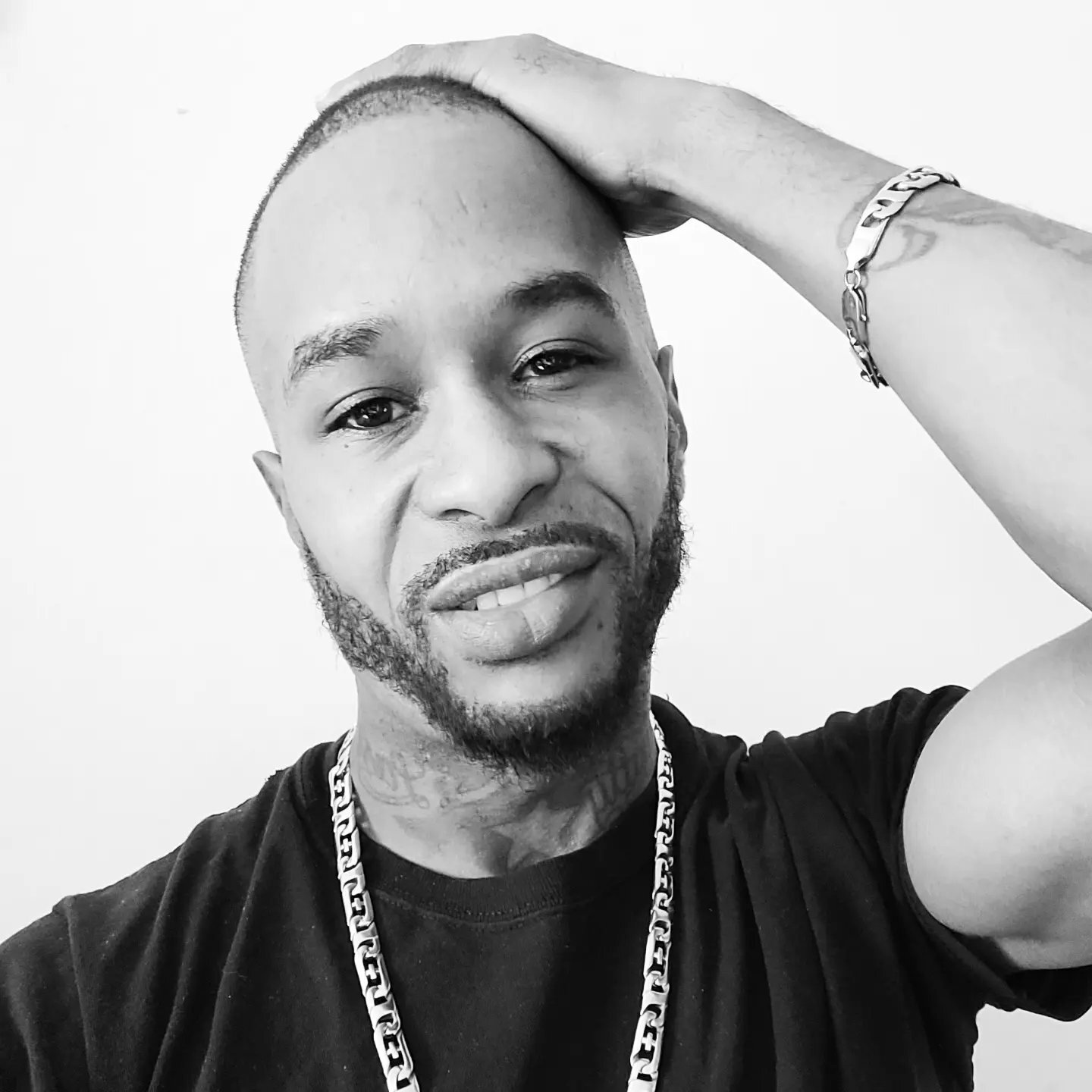 Chris Cardio black-and-white selfie portrait — shaved head, beard, silver chain, black tee, hand on head