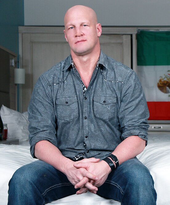 Christian XXX seated portrait — bald, gray paisley western shirt, jeans, between-scenes pose on a studio bedroom set