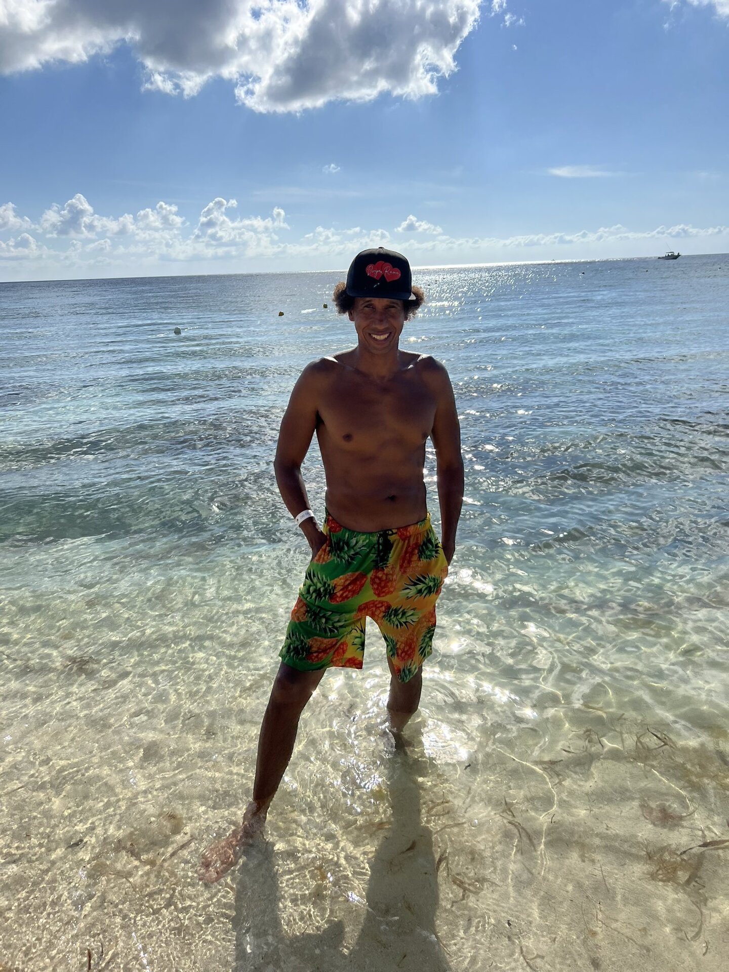 Dvija Rama on a Caribbean beach — branded Dvija Rama baseball cap, pineapple-print board shorts, ankle-deep in clear blue water