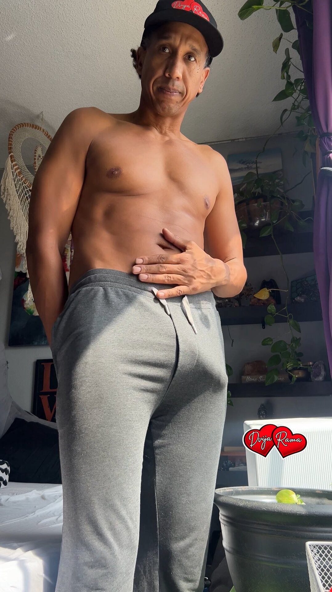 Dvija Rama portrait — shirtless, branded Dvija Rama baseball cap, gray sweatpants, sunlit boho interior with plants and macrame