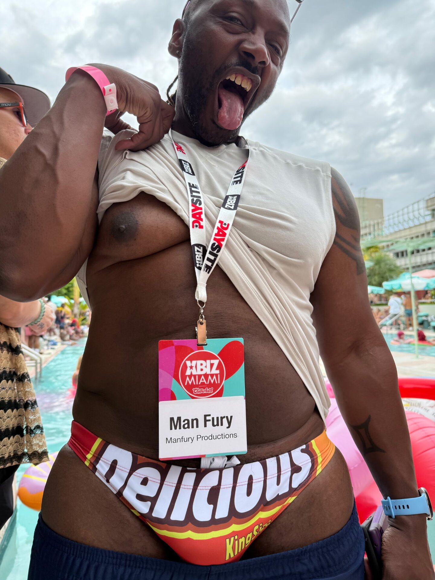 Manfury at XBIZ Miami pool party — bearded with PAYSITE lanyard, 'Man Fury / Manfury Productions' XBIZ badge, candy-print swim brief, playful tongue-out pose