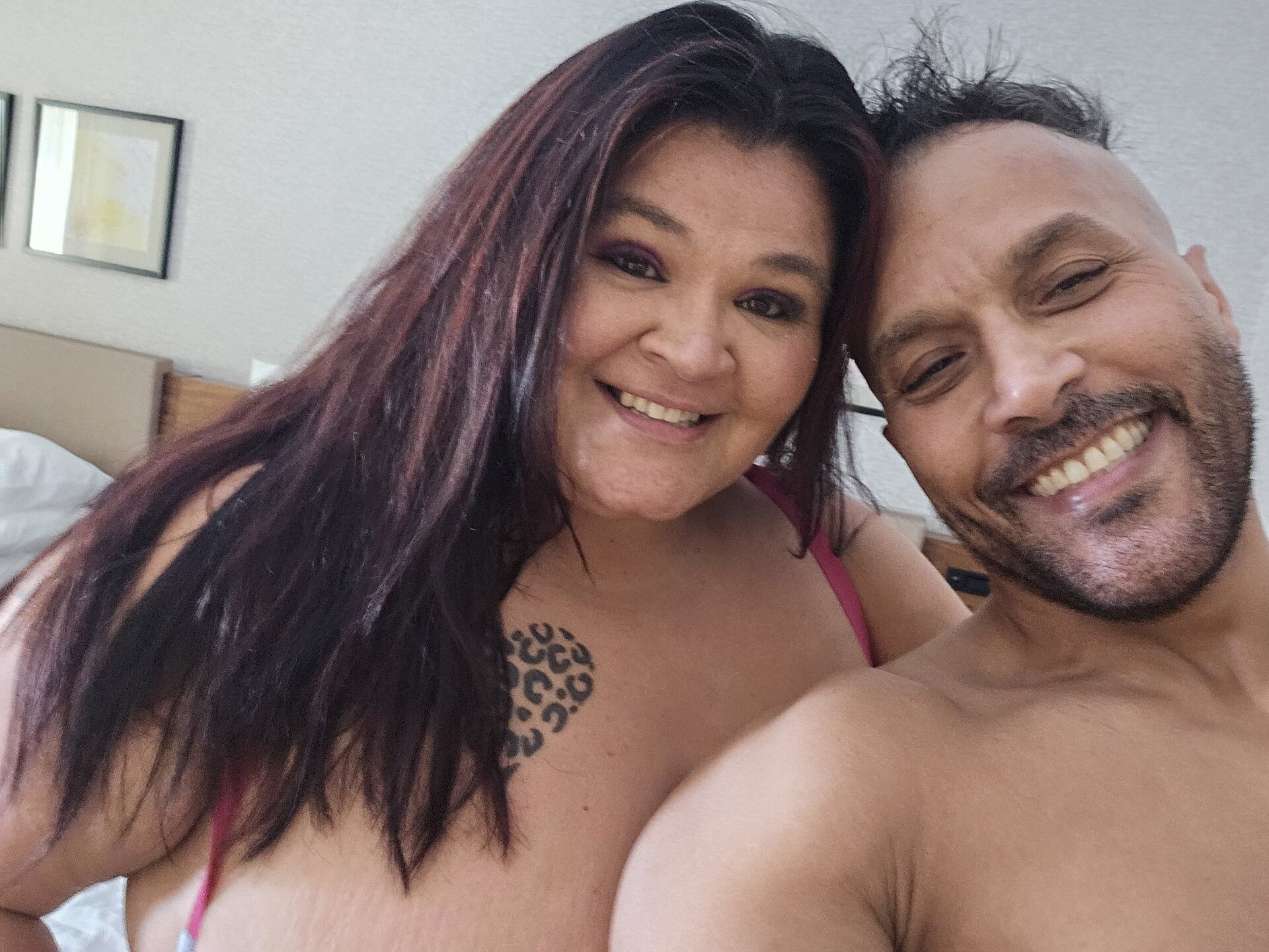 Raven Belle and Chris Getz on-set selfie — hotel bedroom, Raven in pink lingerie, Chris shirtless, both smiling cheek-to-cheek