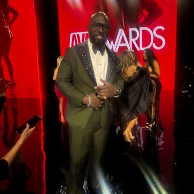 StacXXXs on the AVN Awards red carpet — olive-green satin tuxedo with patterned lapel, in front of the AVN AWARDS backdrop