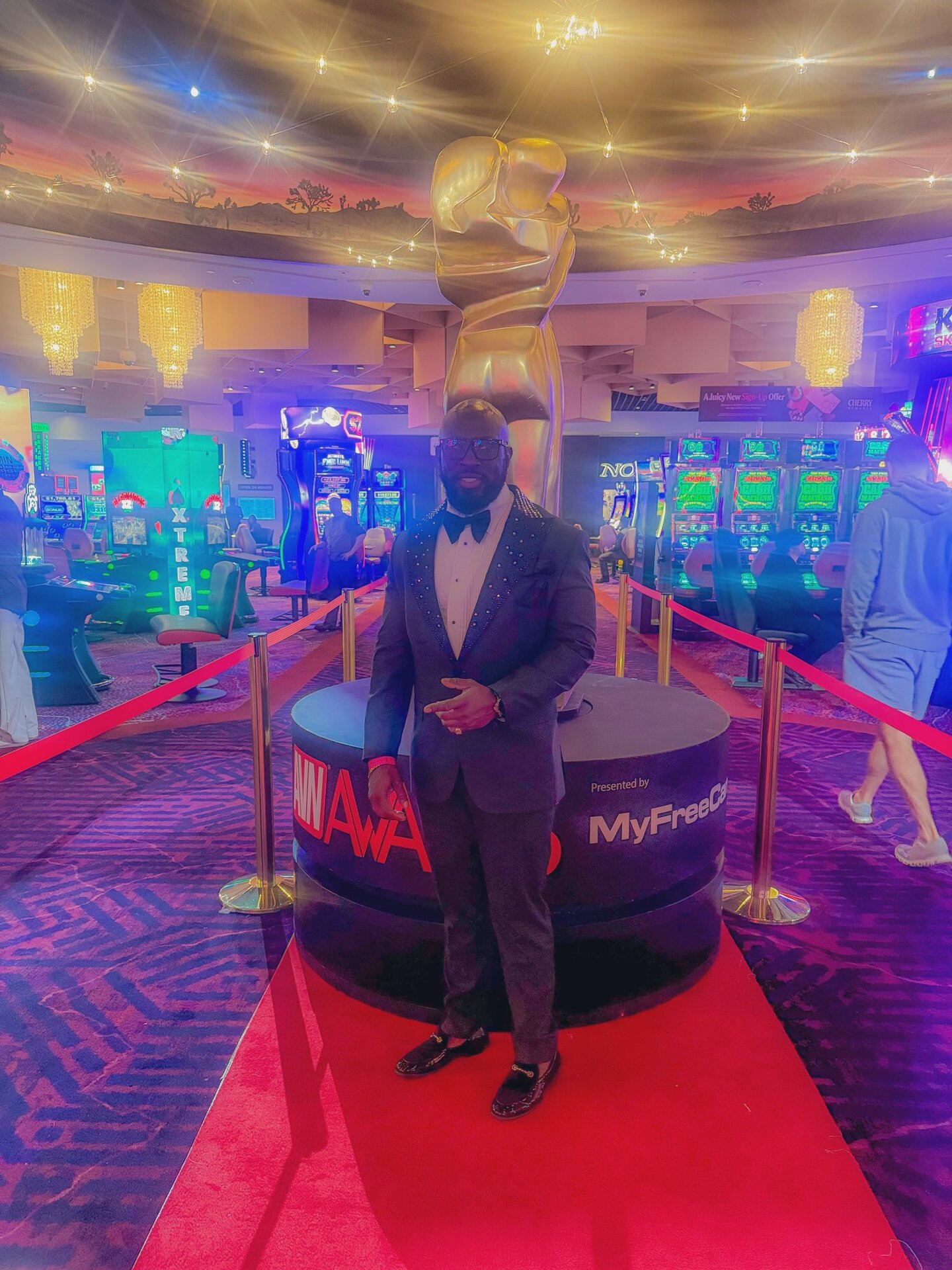 StacXXXs at the AVN Awards — bedazzled black tuxedo with bowtie, standing on the red carpet in front of the giant gold AVN trophy statue