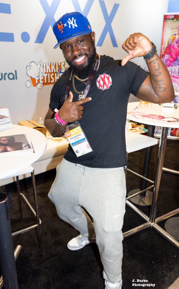 StacXXXs at his expo booth — Mets snapback cap, black 'XXX' tee, gray Nike joggers, AVN exhibitor badge
